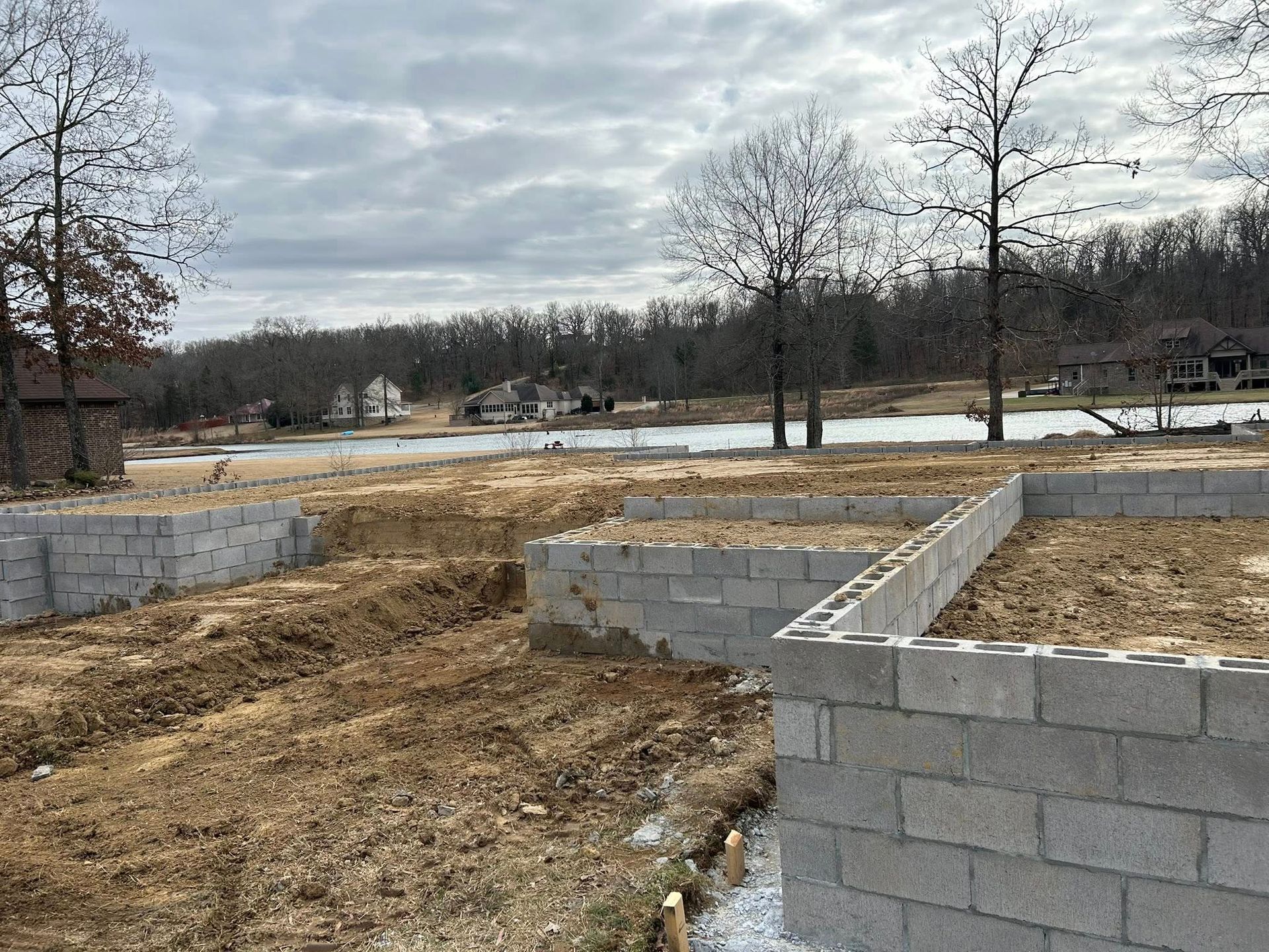 Concrete block foundation walls under construction on a dirt lot overlooking a lake and distant homes.