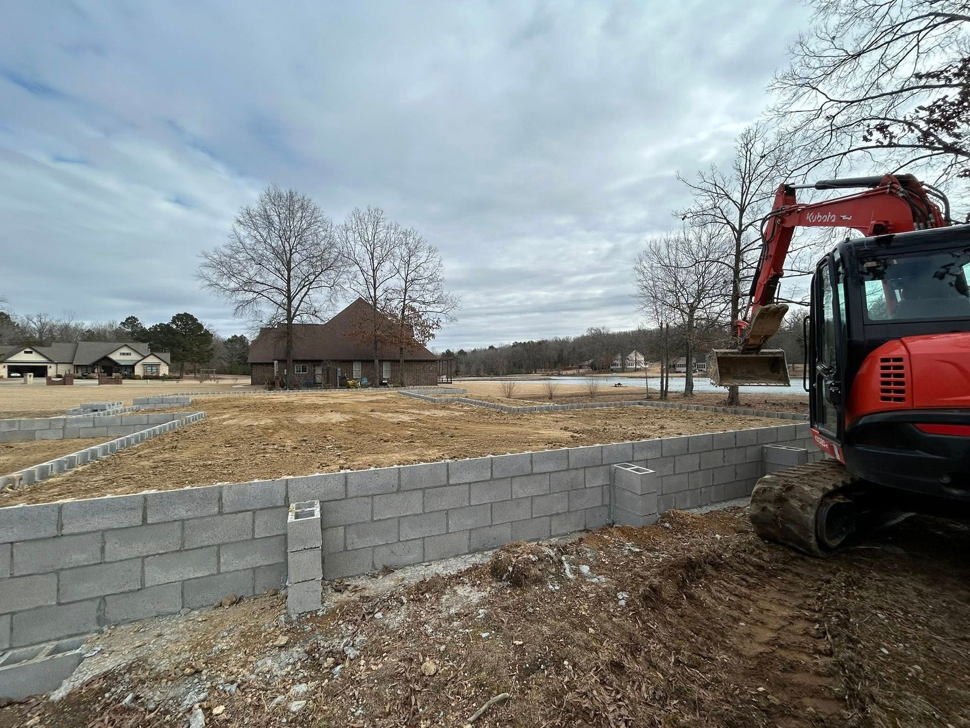 A red excavator sits next to a newly constructed concrete block retaining wall on a dirt lot with a house in the distance.