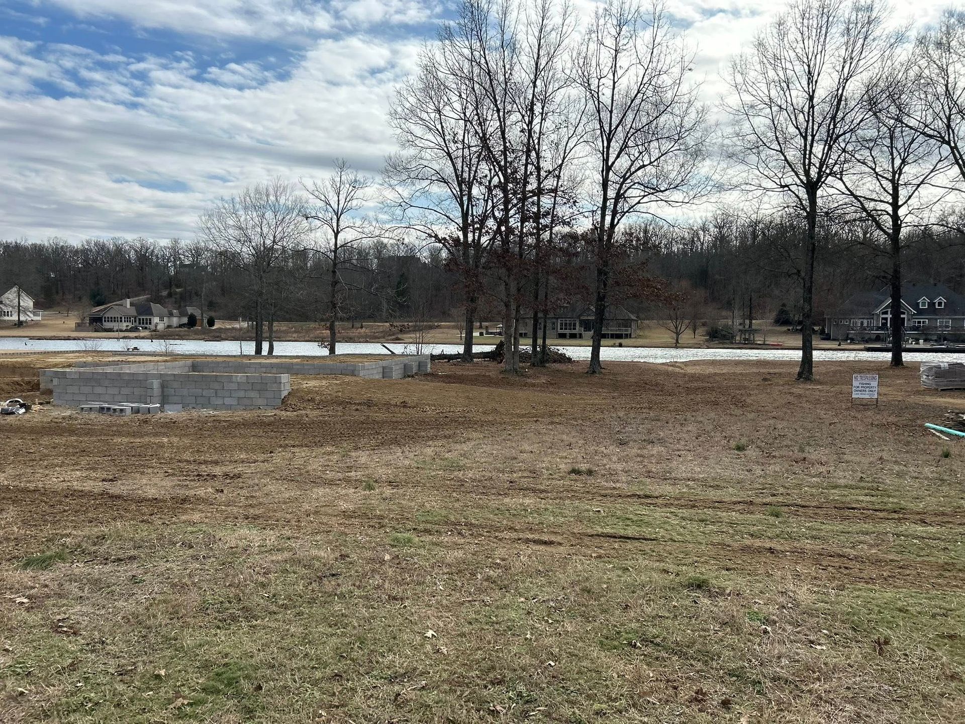 A construction site with a concrete foundation on a grassy lot, surrounded by trees under a cloudy sky.