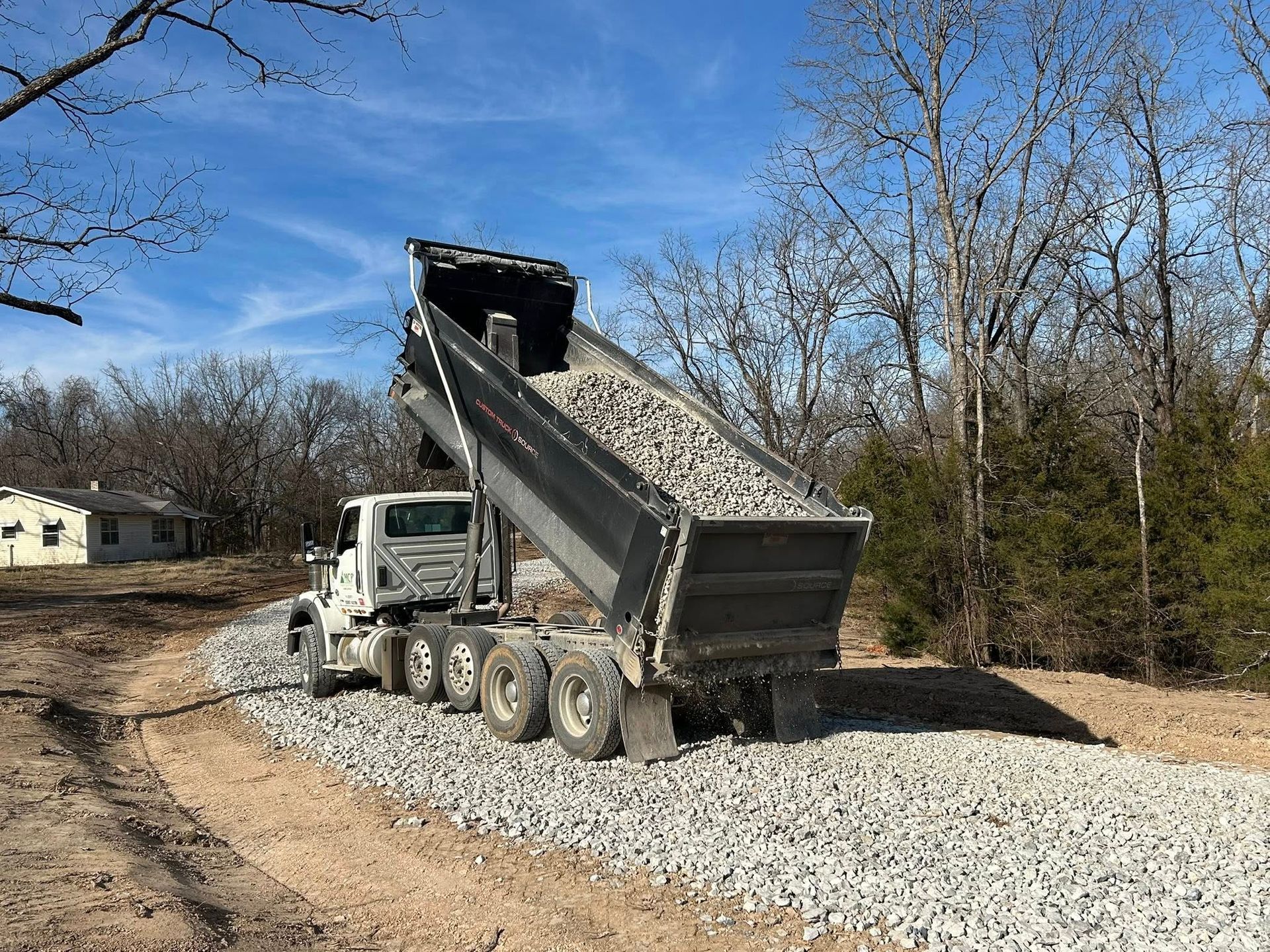 A white dump truck with its bed raised, pouring grey gravel onto a dirt driveway in a wooded area under a blue sky.