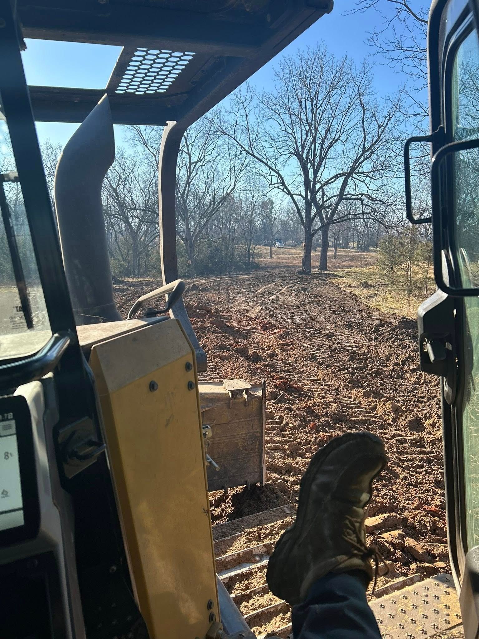 View from inside a heavy equipment cab, showing a person's boot resting on a pedal with an outdoor wooded area ahead.