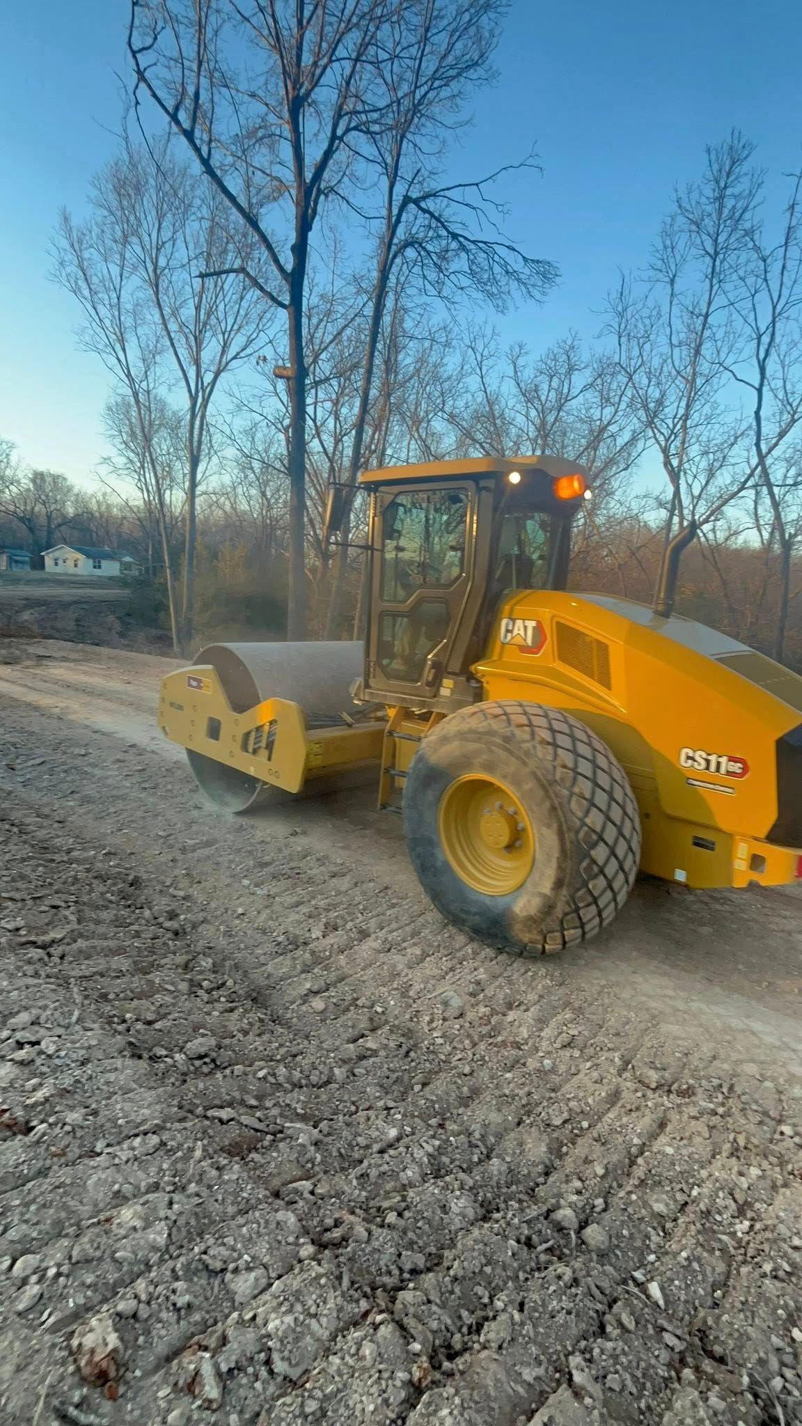 A yellow road roller sits on a dirt construction path in a wooded area under a clear blue sky.