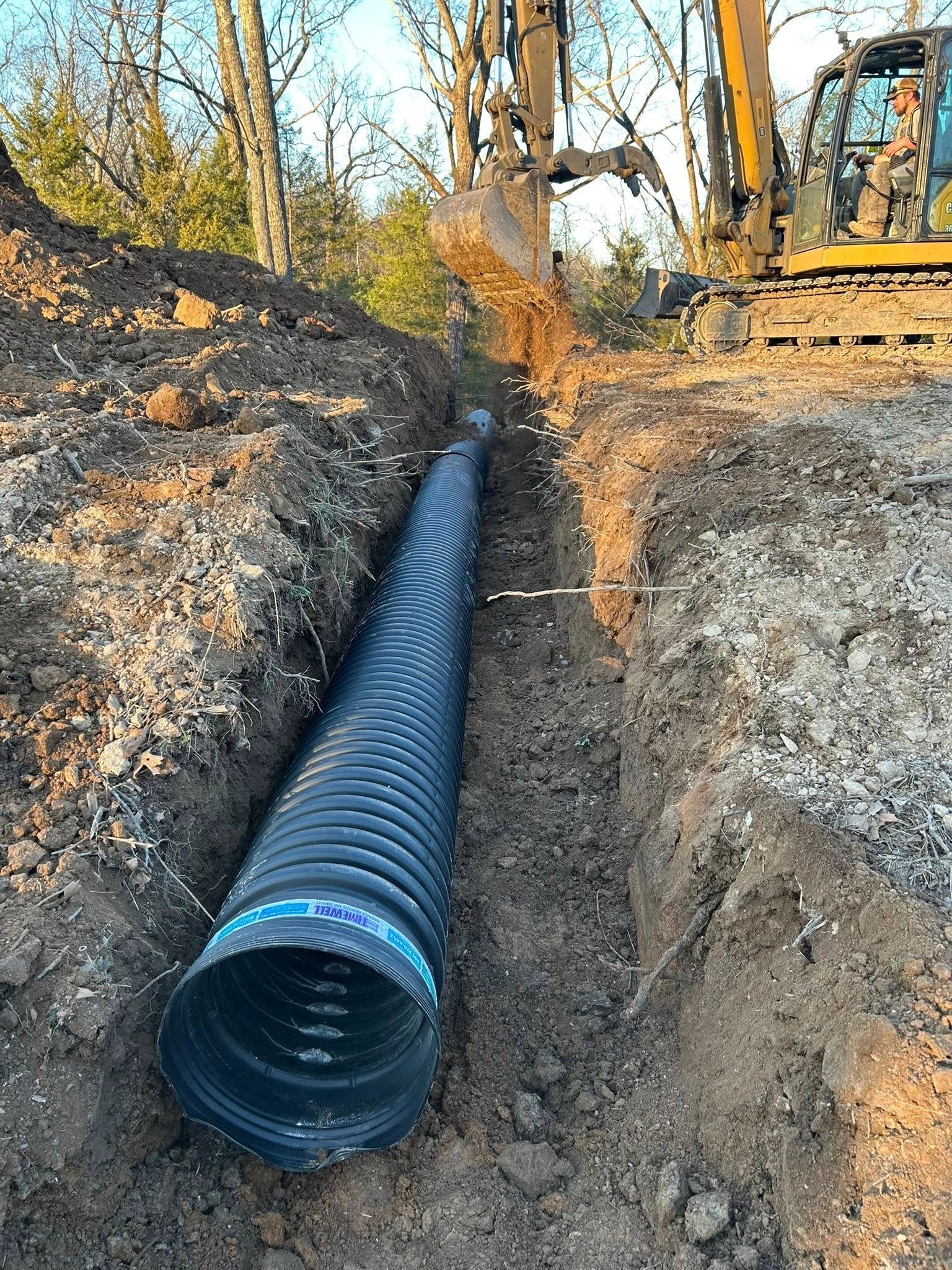 An excavator digs a trench for a black corrugated drainage pipe in a wooded area.