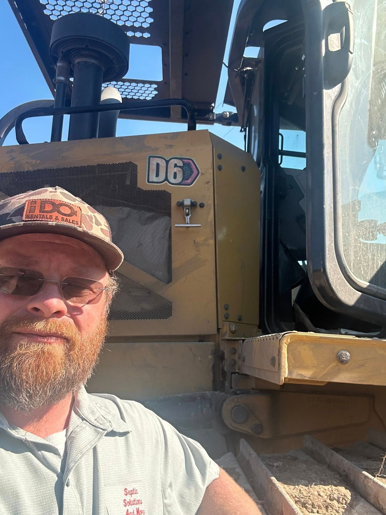 A man with a beard and baseball cap posing for a selfie in front of a yellow Caterpillar D6 bulldozer.