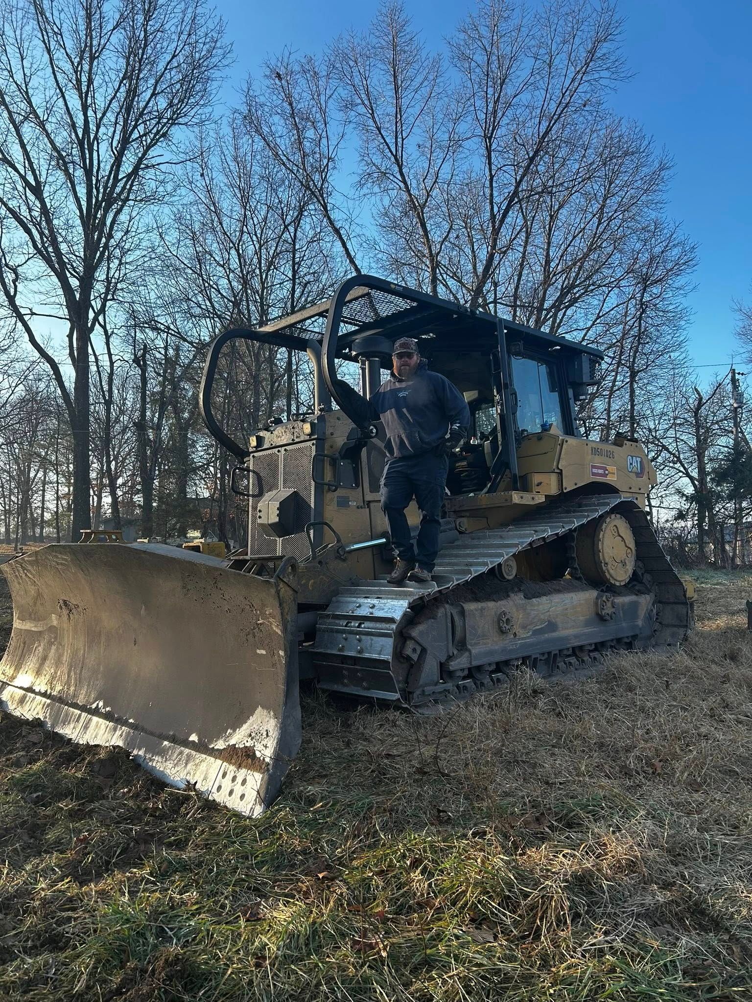 A person stands on the track of a large yellow bulldozer parked in a wooded area under a clear blue sky.