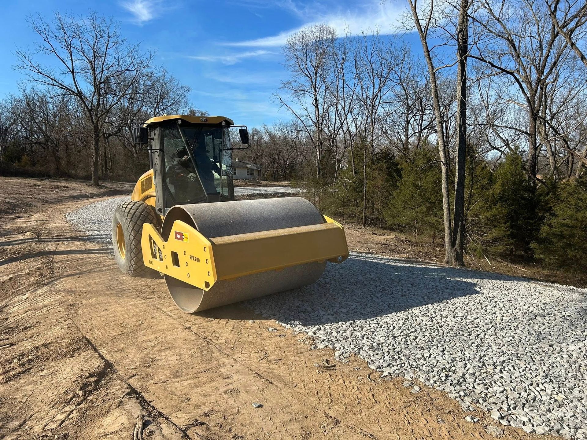 A yellow soil compactor levels fresh gravel on a dirt road in a wooded area under a blue sky.