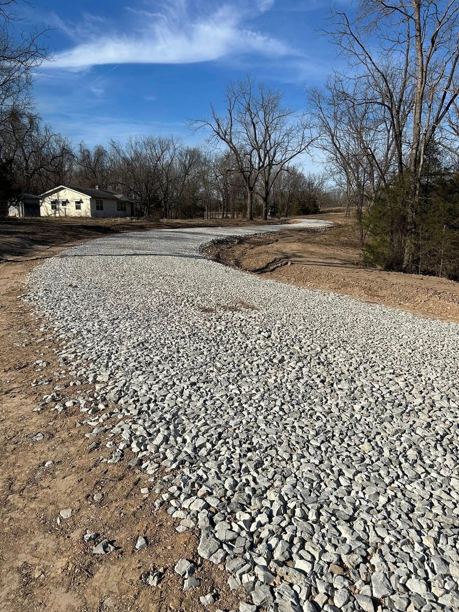 A long gravel driveway curves through a rural, sunlit landscape toward a distant house surrounded by bare trees.