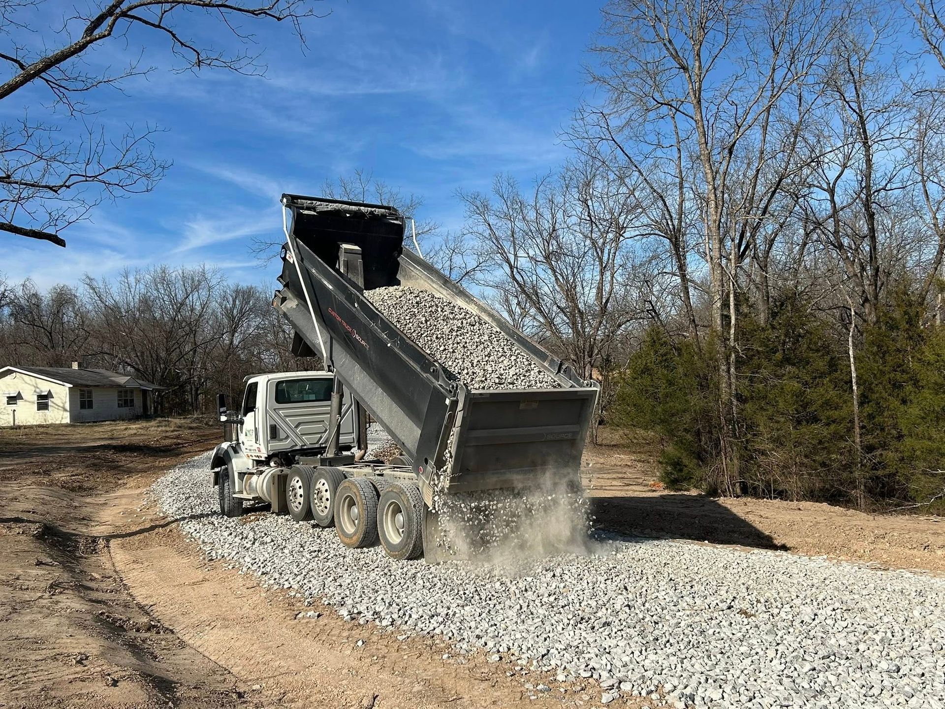 A dump truck in a wooded area deposits gravel onto a dirt path under a clear blue sky.