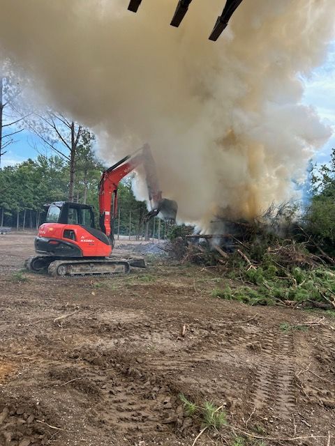 An orange excavator sits beside a large pile of brush and debris emitting a thick plume of smoke in a cleared field.
