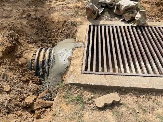 A corrugated pipe connects to a rectangular metal storm drain grate, with wet concrete sealing the joint in a dirt pit.
