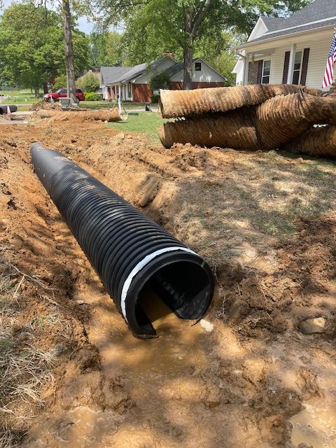A black corrugated drainage pipe sits in a muddy trench in front of a residential house.