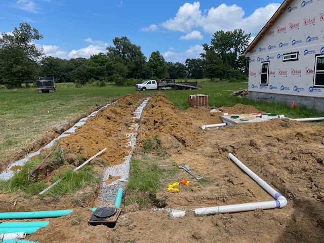 Construction site showing a house foundation with exposed pipes, an open septic trench, and a truck in a grassy field.