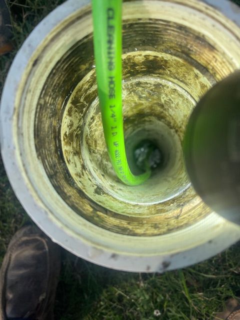 A bright green hose snakes into a vertical, light-colored pipe, with a person's shoe visible at the bottom of the frame.