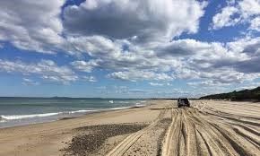 A car is driving down a sandy beach next to the ocean.