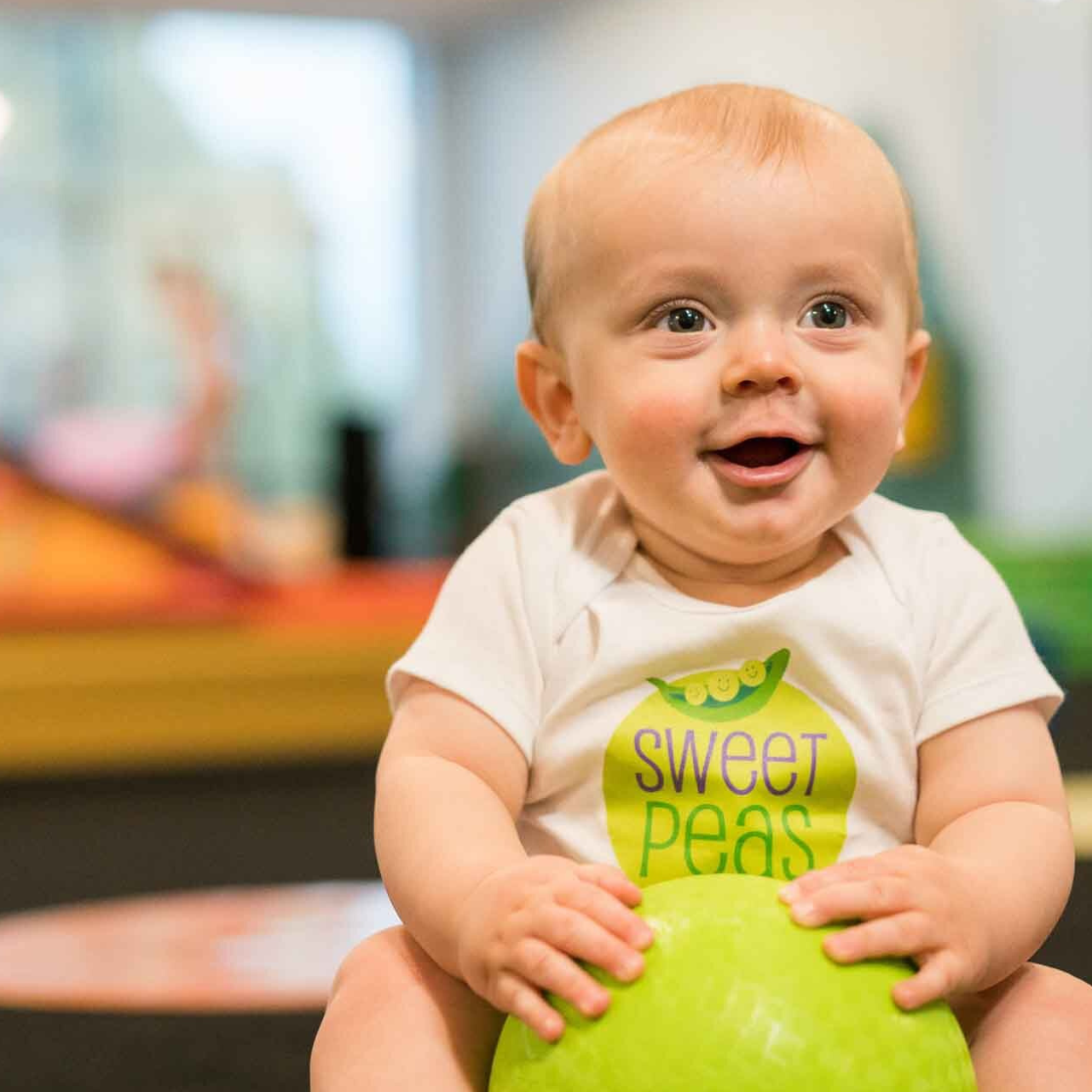 A baby wearing a sweet peas shirt is sitting on a green ball.