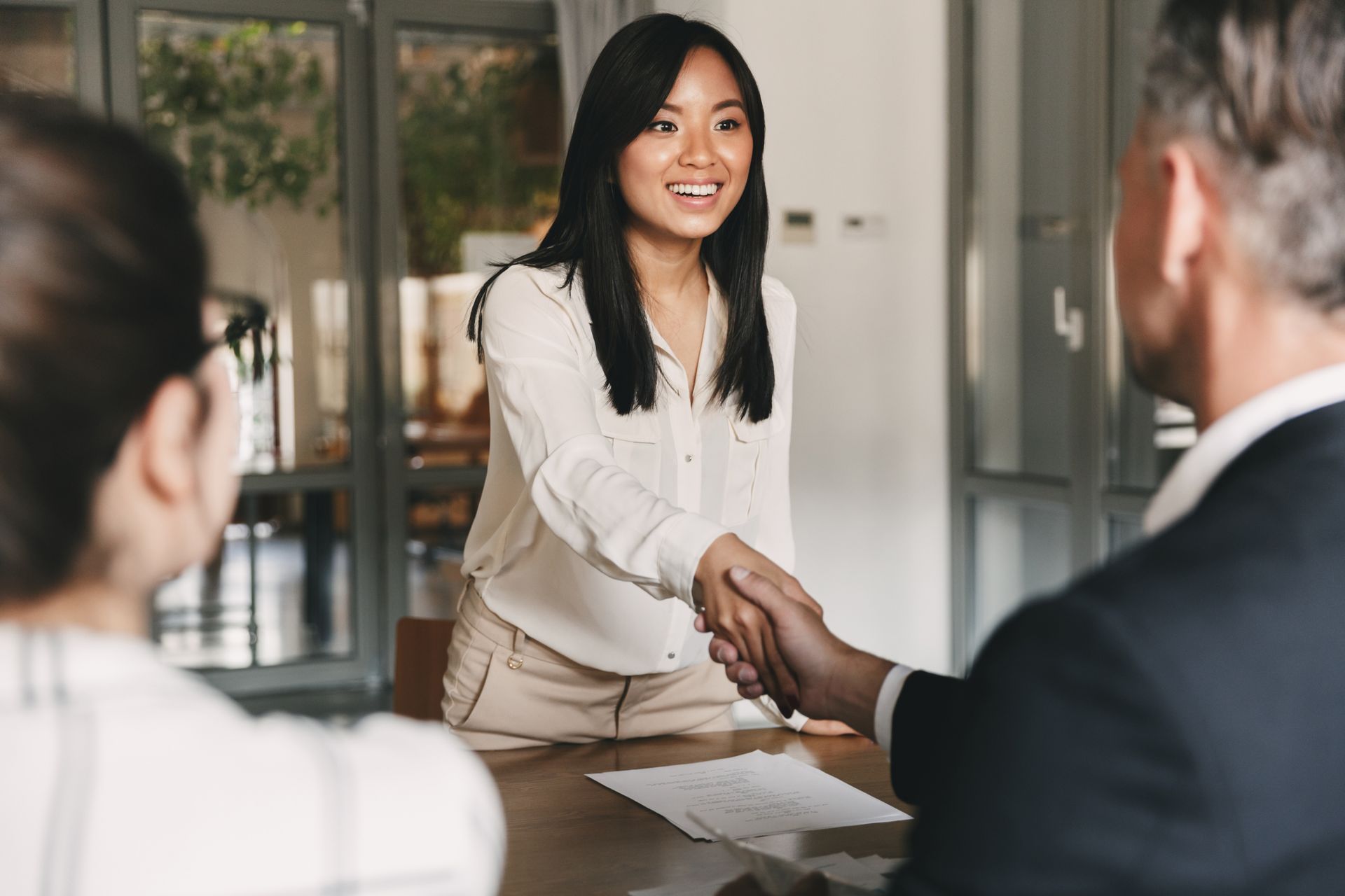 Woman in white shirt shaking hands across a table in a bright office meeting.