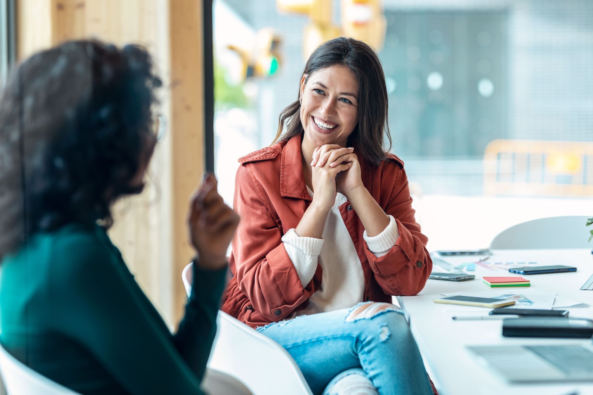 Two people talking at a bright office table, one smiling in a red jacket