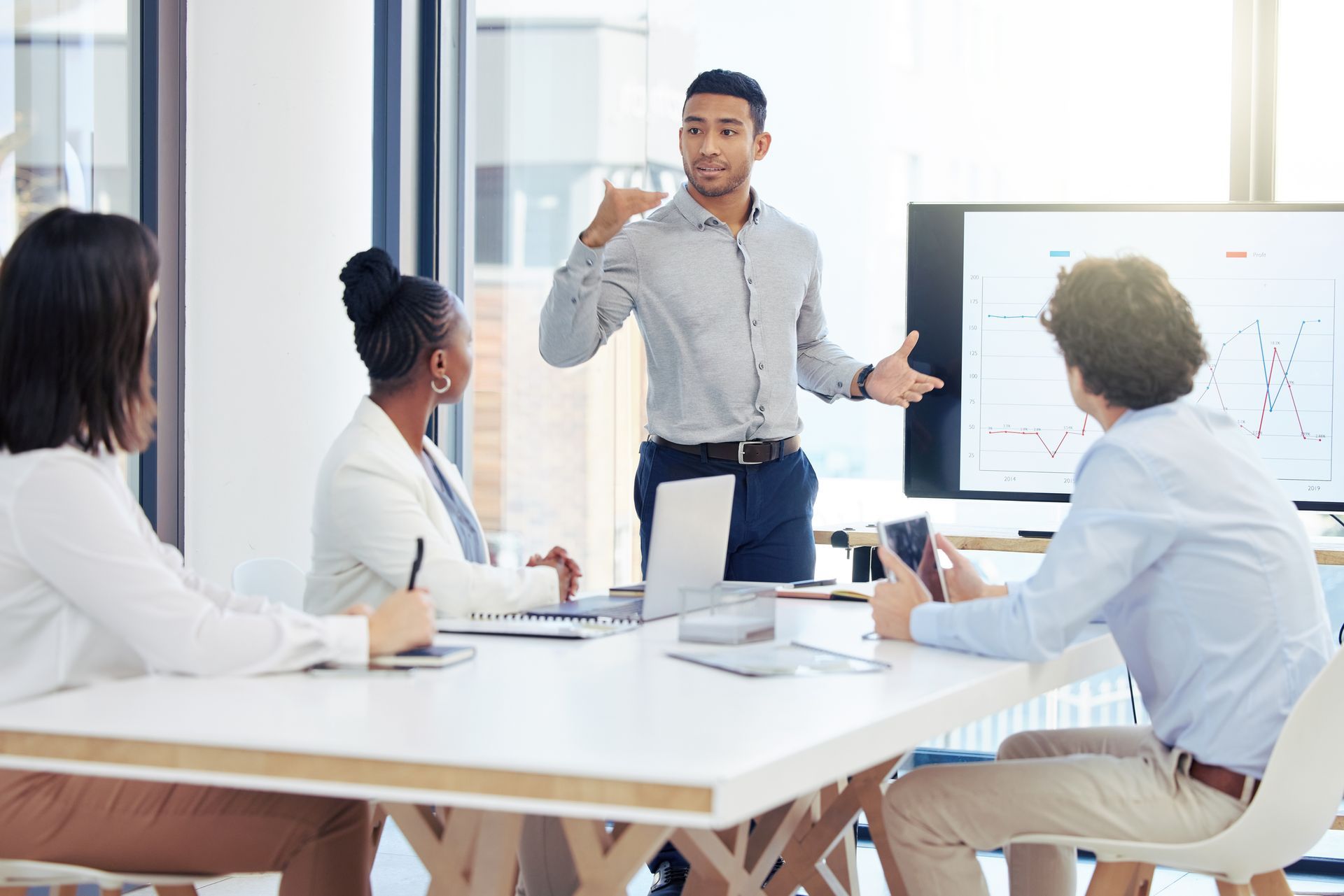 Business meeting with four people around a conference table, listening to a presenter near a screen.