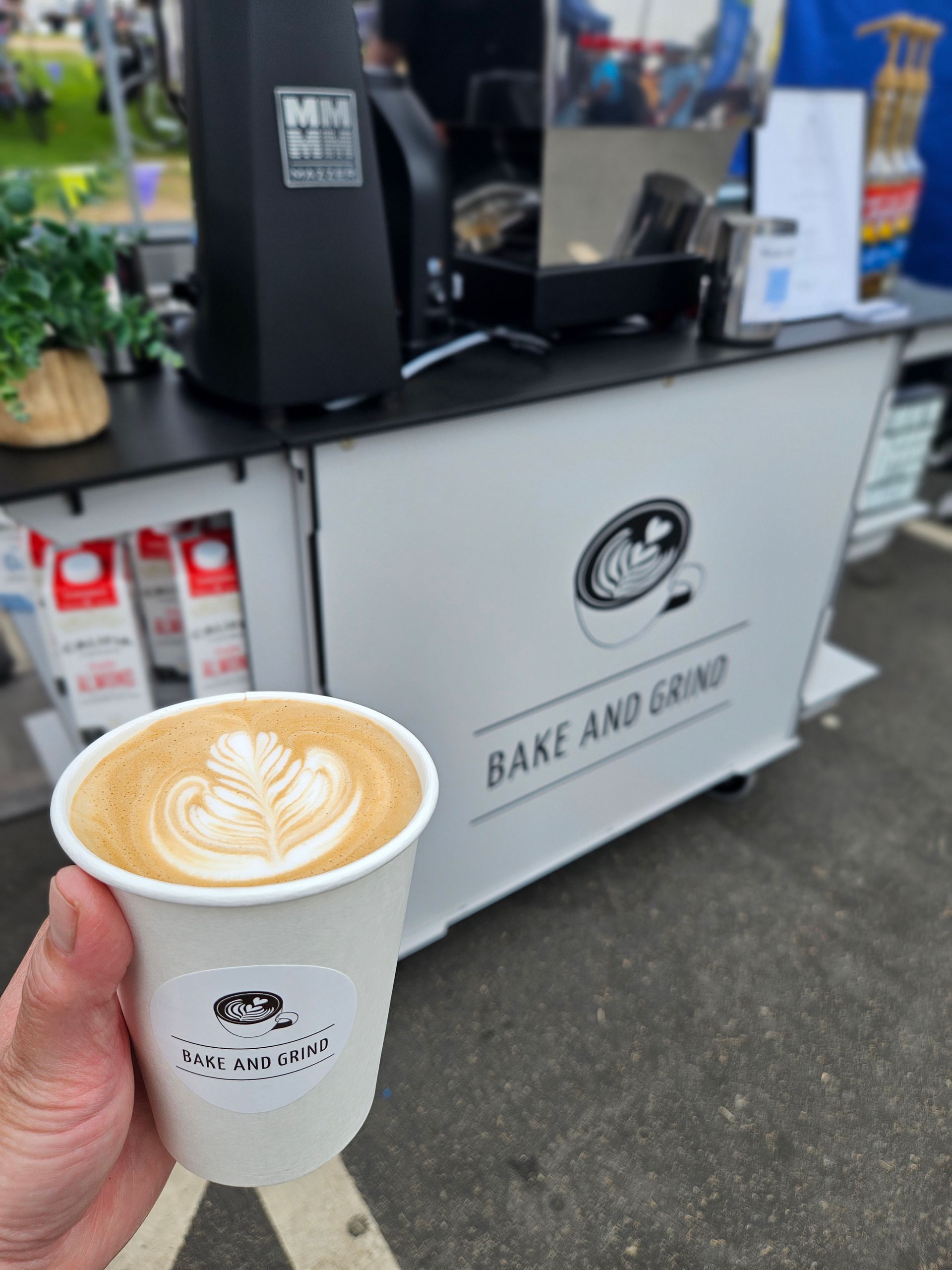 Hand holding a latte in front of a coffee cart with a “Bake and Grind” sign
