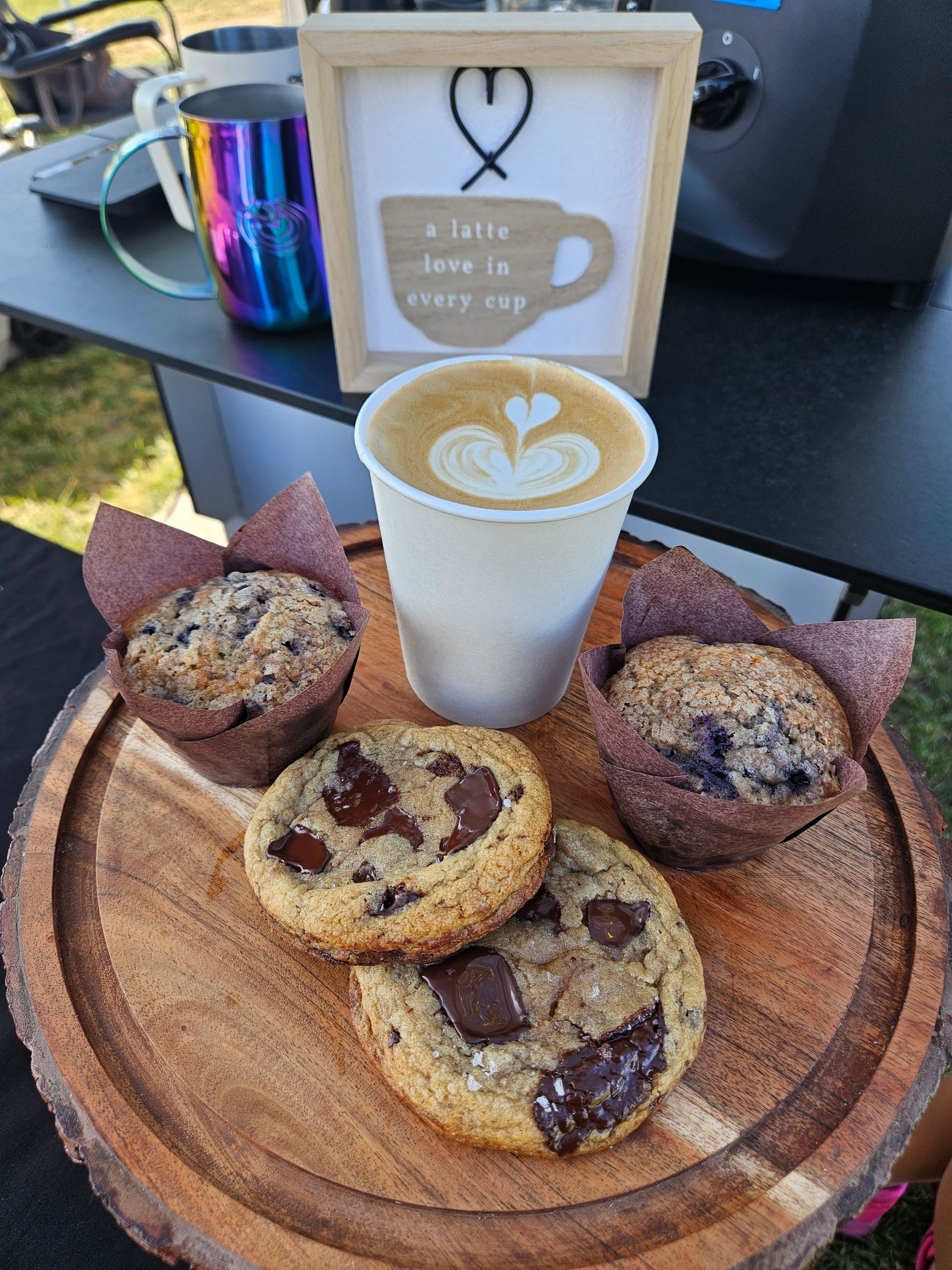 Coffee with four chocolate-chip muffins on a wooden board at an outdoor table.