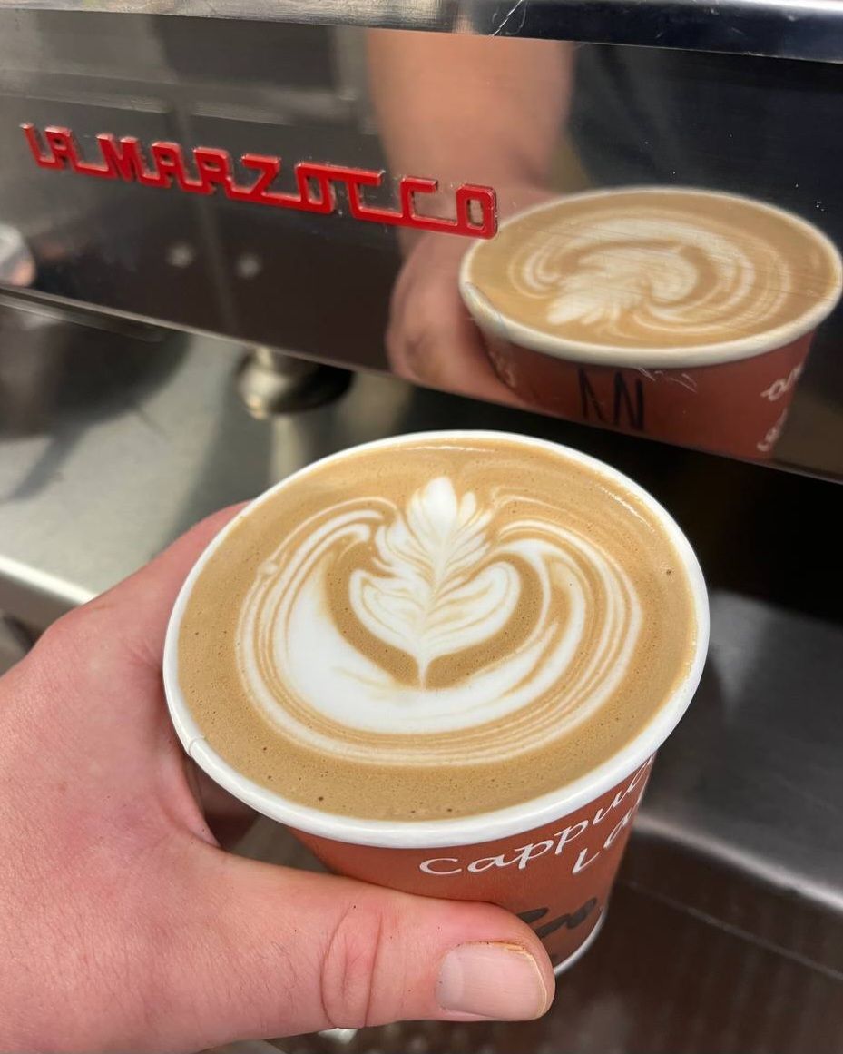 Hand holding a small takeaway cup of latte with heart latte art beside a shiny espresso machine.