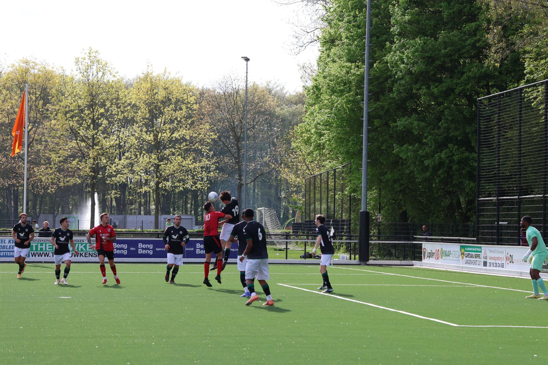 Voetballers strijden om een ​​hoge bal vlakbij het doel op een buitenveld in de felle zon.