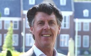 Een lachende man in een wit shirt staat buiten voor bakstenen gebouwen.