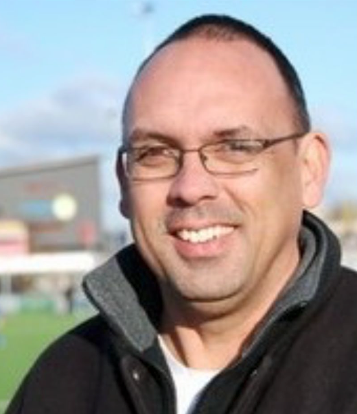 Lachende man met bril en donkere jas buiten op een zonnige dag.