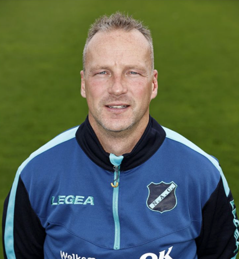 Man in een blauw sportjack met embleem, staand tegen een groene veldachtergrond.