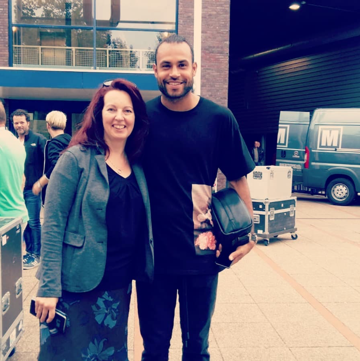 Twee mensen glimlachen buiten, dicht bij elkaar staand in de buurt van een gebouw en een geparkeerde bestelwagen.