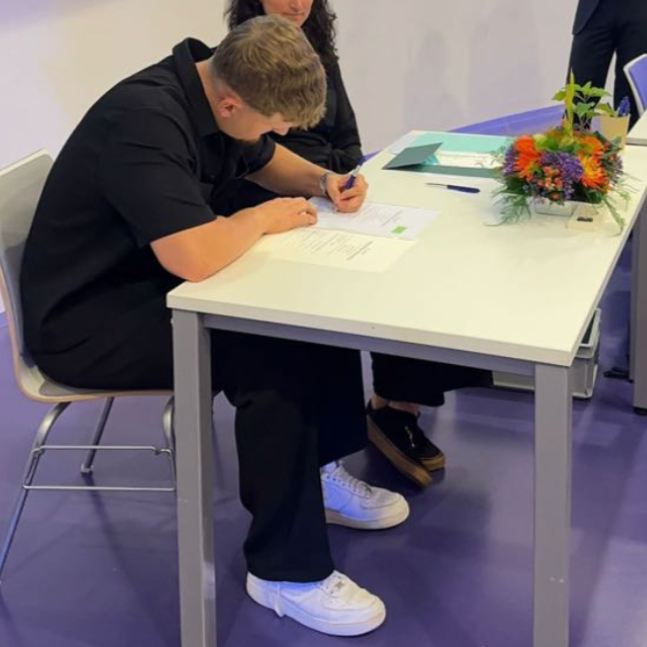 Een persoon zit aan een witte tafel en schrijft op papier, met bloemen en een turquoise map in de buurt.