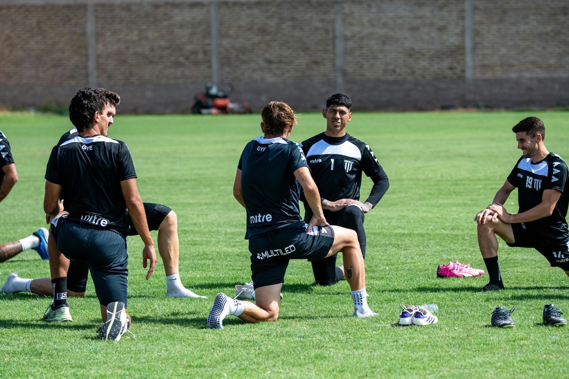 Atleten knielen op een grasveld tijdens een voetbaltraining in de buitenlucht, gekleed in donkere trainingskleding.
