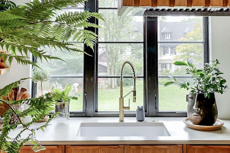 Kitchen with gold faucet, sink, black-framed window overlooking a yard; plants on counter and in the window.