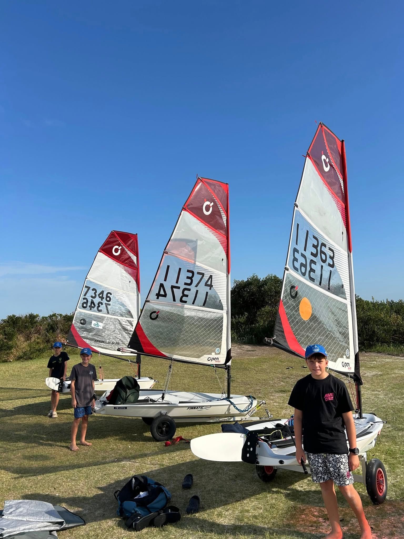 Three Sailboats With Red and White Sails on a Grassy Field Under a Blue Sky With Three Children — Gosford Sailing Club in Gosford, NSW