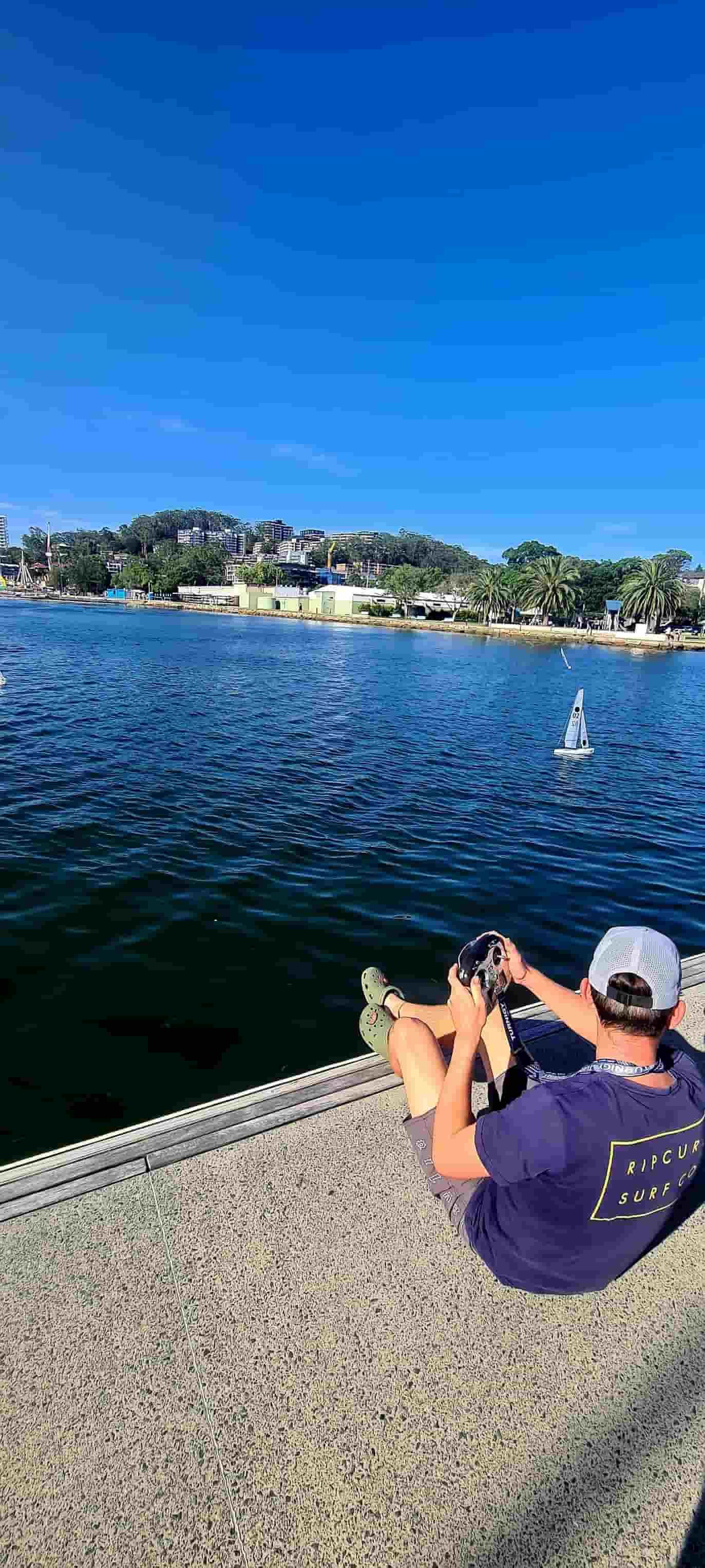 A Man is Sitting on a Dock Taking a Picture of a Boat in the Water — Gosford Sailing Club in Gosford, NSW
