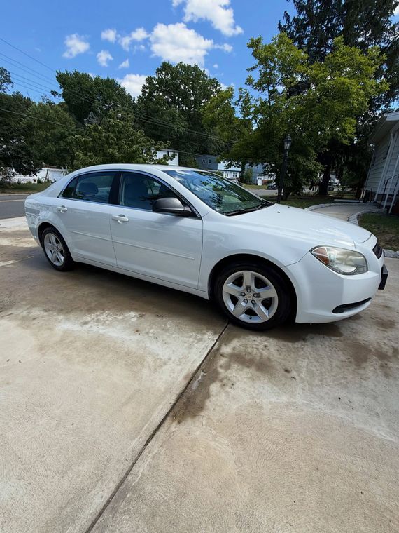 White sedan parked on a wet driveway beside trees and a house under a blue sky.