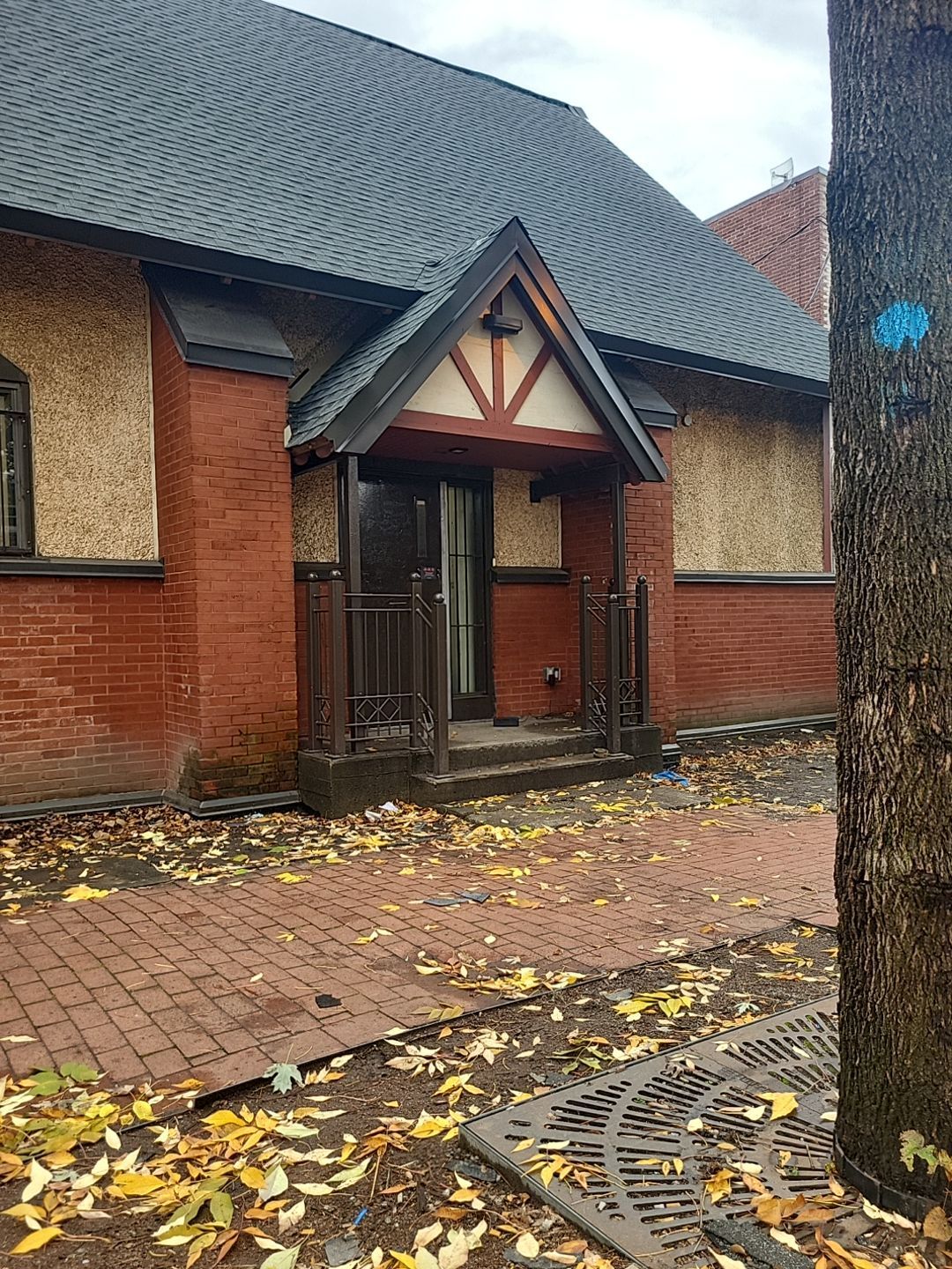 Bâtiment en briques rouges avec une entrée de style Tudor sous un toit noir. Feuilles mortes sur le chemin et le sol en briques.