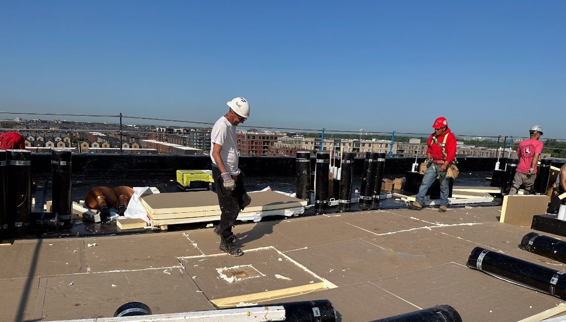 Des ouvriers du bâtiment sur un toit portant des casques de sécurité ; l'un marche, d'autres travaillent avec des matériaux.