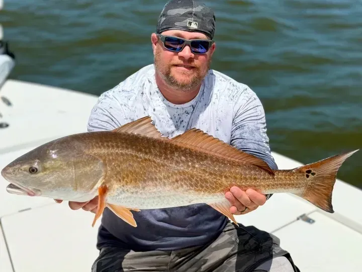 Man holding a large redfish on a boat; sunlight, blue water.