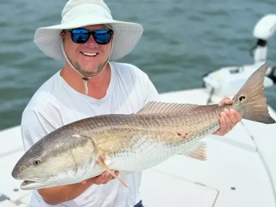 Man smiling, holding a redfish on a boat, wearing sunglasses and a hat.