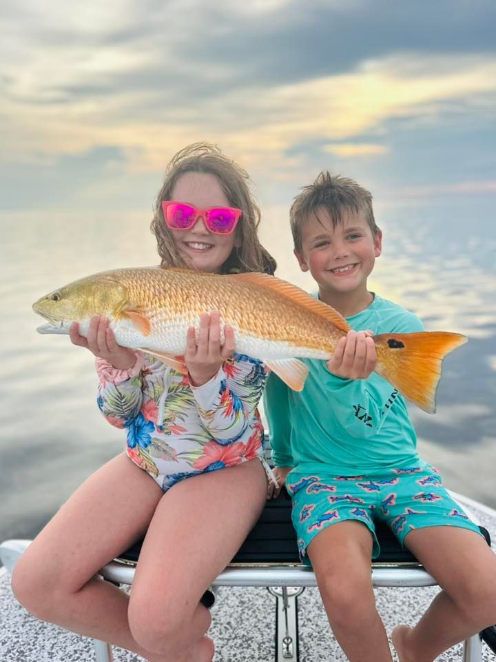 Two children holding a large redfish on a boat; smiling with a sunset background.