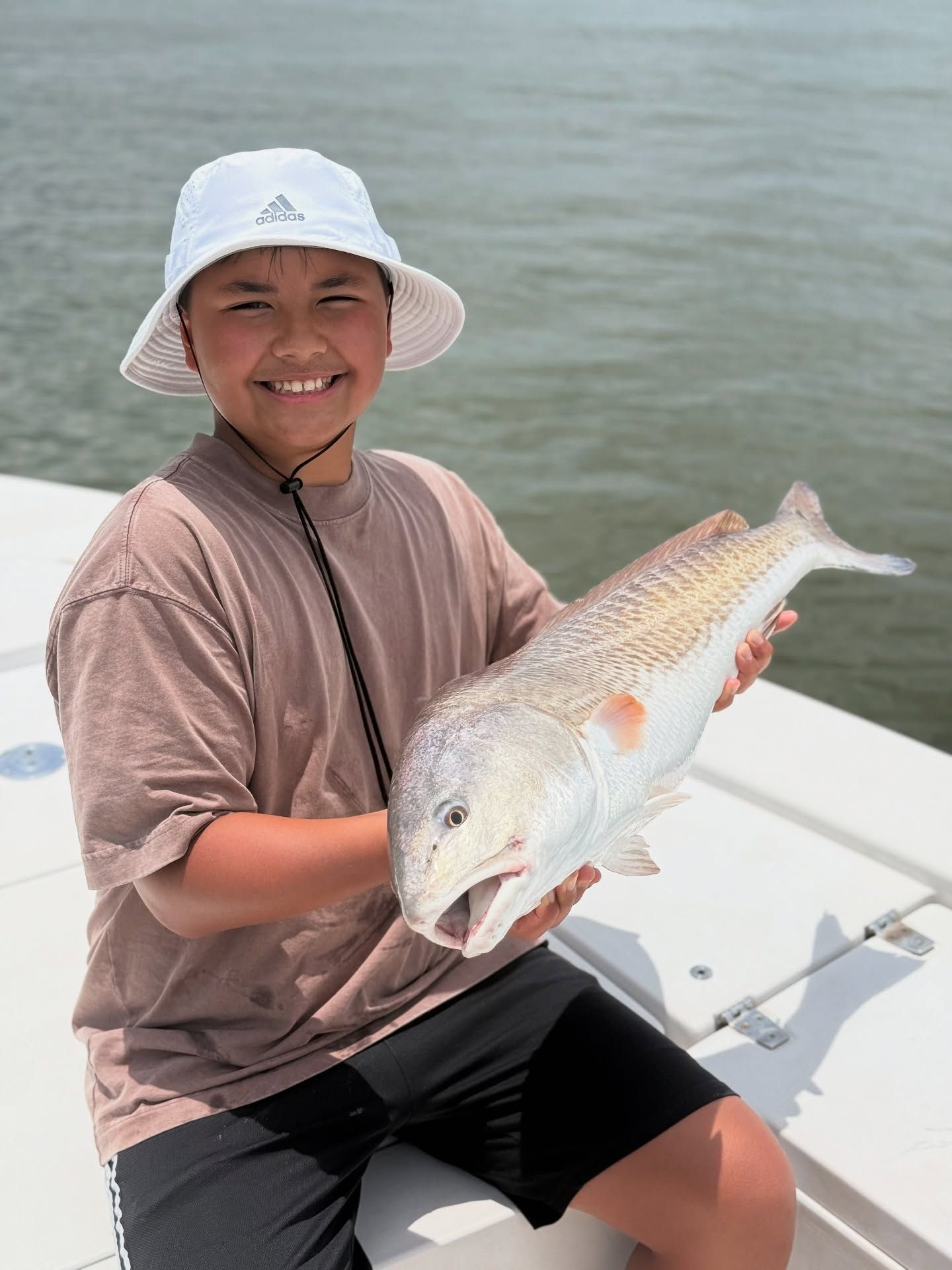 Boy holding a redfish on a boat, smiling with a white hat in a water setting.