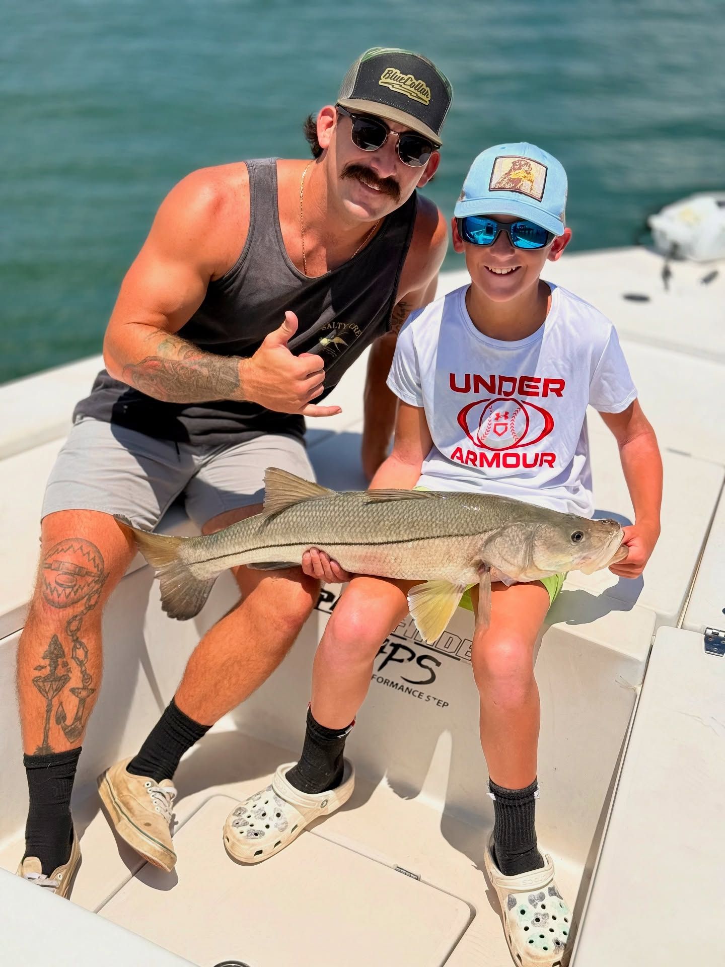 Man and child on a boat holding a fish, sunny day. The man has a mustache and tattoos.