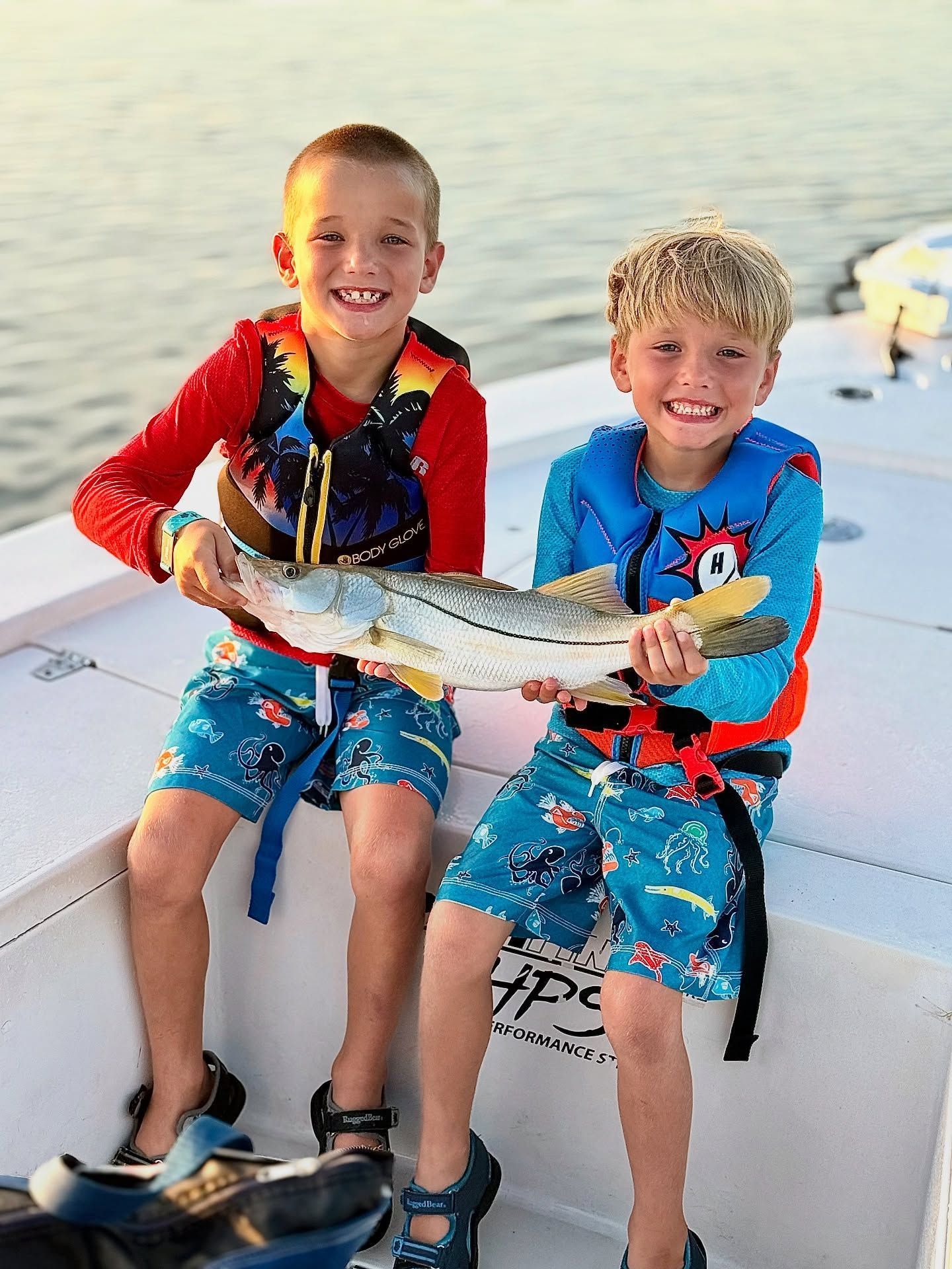 Two boys on a boat holding a fish, smiling. They wear life vests and swim trunks.