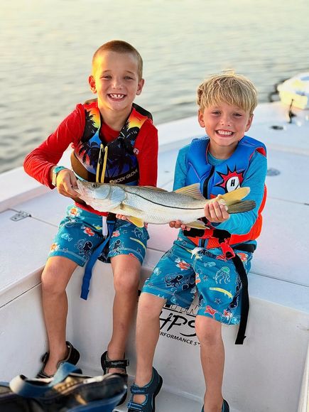Two boys on a boat holding a fish, smiling. They wear life vests and swim trunks.