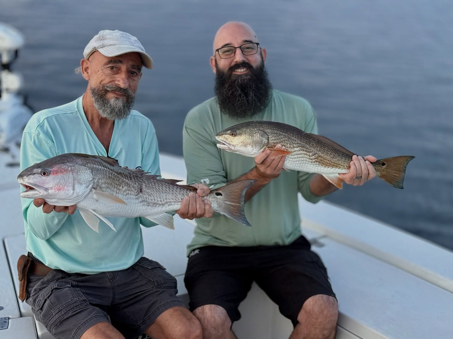 Two men on a boat hold up redfish they have caught. Calm water in the background.