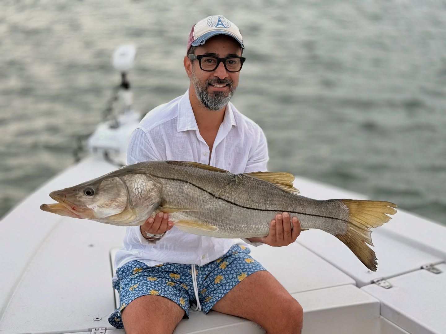Man on boat holding up a large fish, smiling. Overcast sky.
