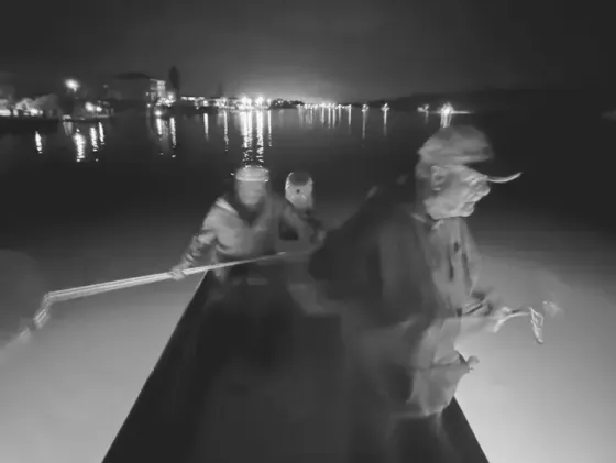 Three people in a boat on dark water at night; city lights in the distance.