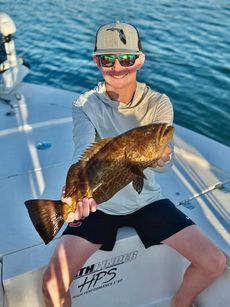 Person on a boat holding a large fish, wearing sunglasses, Florida hat, and shorts. Blue water in background.