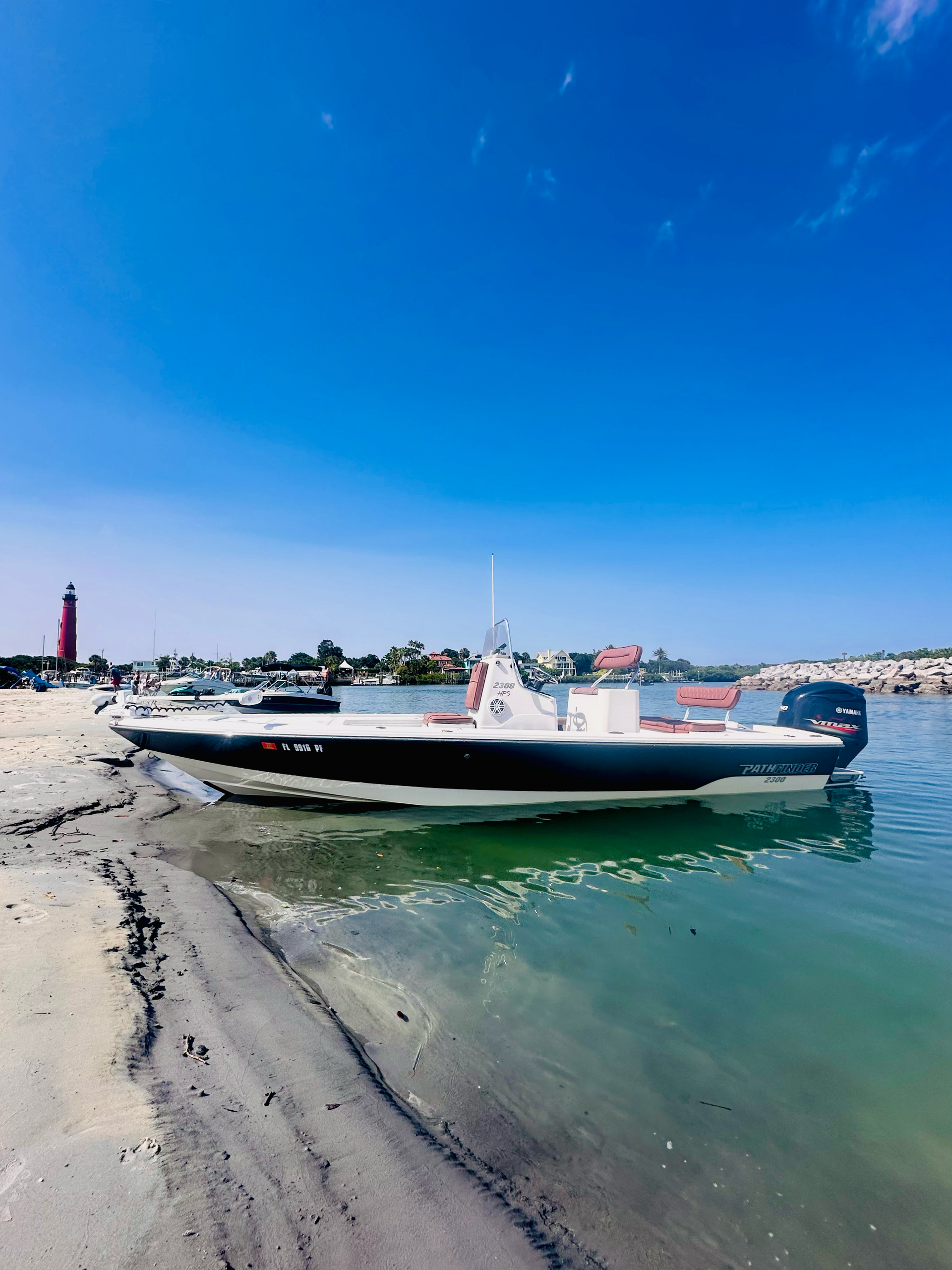 Boat beached on sandy shore under a bright blue sky, with a red lighthouse in the background.